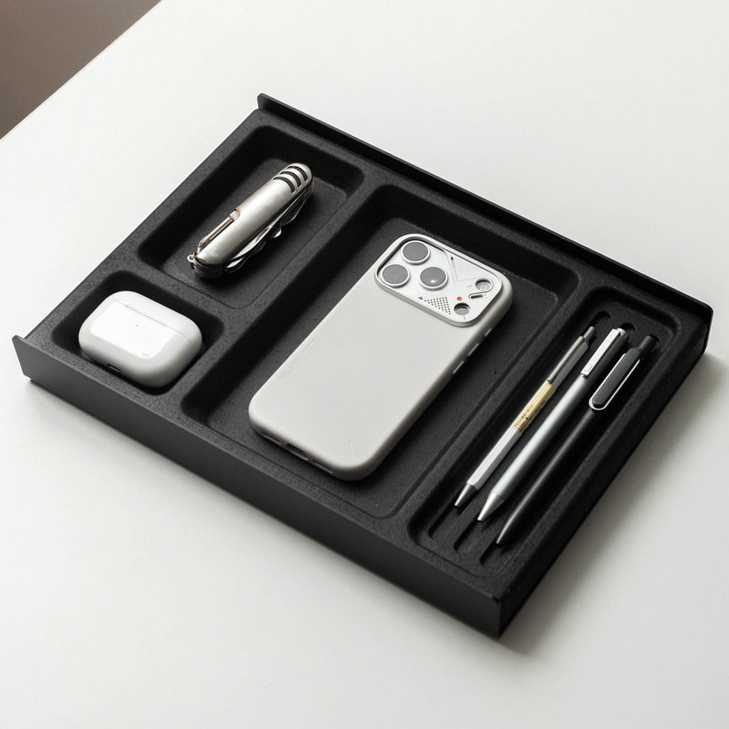 A black cork and aluminium organizer tray holding a white phone case, a white earbud case, a silver pen, and a metallic accessory, photographed on a white surface. The image includes the text 'Seamless' at the top and small footer text at the bottom.